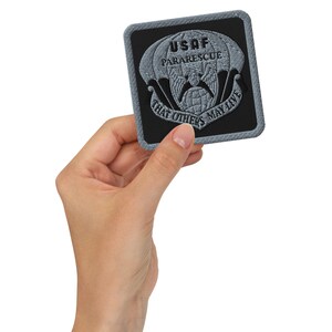Puede incluir: Un parche cuadrado con fondo negro y diseño bordado gris. El diseño presenta un paracaídas, un globo terráqueo y el texto "USAF Pararescue" y "That Others May Live". El parche tiene un borde tipo denim.