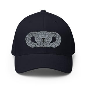 Op de afbeelding: Marineblauwe baseballcap met een zilveren embleem met een wereldbol, vleugels en laurierbladeren. De cap heeft een gebogen klep en een gestructureerde pasvorm. Het embleem is gecentreerd op de voorkant.