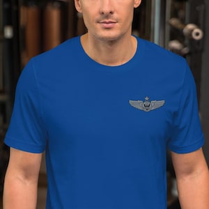 May include: A royal blue t-shirt featuring a silver-colored emblem of wings, a star, and a circular design. The emblem is located on the upper left chest area of the shirt. The shirt is made of a soft material.