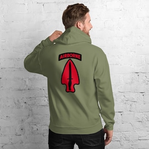 May include: Olive green hoodie with the word "AIRBORNE" above a red arrowhead graphic. The graphic features a black dagger. The hoodie has a drawstring hood and long sleeves. The person in the image is wearing the hoodie.