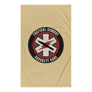Könnte beinhalten: Beige Banner mit einem kreisförmigen Emblem mit dem Text "TACTICAL COMBAT CASUALTY CARE". Das Emblem zeigt ein schwarzes Messer über einem roten Blitz und ein weißes Lebenssternsymbol, alles in einem schwarzen und roten Kreis.