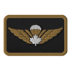 May include: Embroidered patch featuring a black background with a gold border. The design includes gold wings, a white maple leaf, and a parachute with white and gold segments. The patch is rectangular with rounded corners.