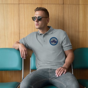 Może przedstawiać: Szara koszulka polo z niebieskim kółkiem wyszytym na lewej piersi. Koło zawiera stylizowane zdjęcie statku kosmicznego.