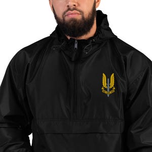Puede incluir: Chaqueta negra con capucha y cierre de media cremallera. La chaqueta presenta un emblema amarillo y negro en el pecho con el texto "Who Dares Wins". Tiene capucha con cordón y un bolsillo delantero. Prenda de vestir informal.