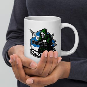 Puede incluir: Taza de cerámica blanca con un gráfico de una parca con una capa con capucha verde sosteniendo una ametralladora. El diseño incluye un emblema azul y blanco con la palabra "GHOST" en un óvalo negro. La taza tiene un asa grande y curvada.