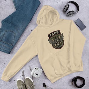 Pode incluir: Um moletom creme com um bolso frontal e um capuz. O moletom apresenta um patch com o texto "TACP", uma caveira com asas e as palavras "CLEARED HOT". Calças de ganga azuis, auscultadores e um smartphone também são visíveis.