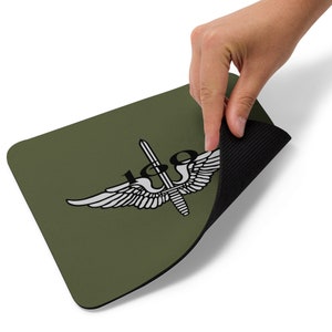 May include: A green mouse pad with a white and black military insignia featuring a sword and wings. The insignia has the text "180" on it. The mouse pad has a black rubber backing.