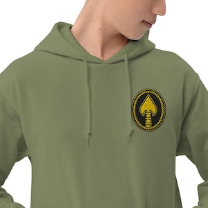 Pode incluir: Camisola com capuz verde-oliva com um patch bordado a ouro e preto no peito. O patch apresenta um design de espadas douradas dentro de um círculo preto, rodeado por uma borda dourada em forma de corda. A camisola tem um capuz com cordão.
