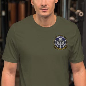 Könnte beinhalten: Olivgrünes T-Shirt mit einem kreisförmigen Emblem mit der Aufschrift "TASK FORCE PHANTOM TALON". Das Emblem zeigt einen Totenkopf, ein Schwert und Lorbeerblätter. Das T-Shirt ist aus weichem Material.