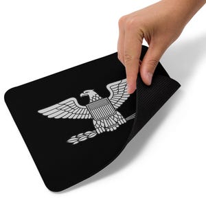 Op de afbeelding: Zwart rechthoekig muismat met een wit adembleem, gespreide vleugels, schild en lauriertak. De muismat wordt opgetild en toont een zwarte, getextureerde onderkant.