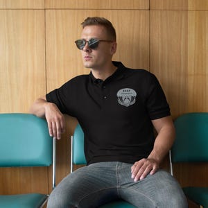 Puede incluir: Polo negro con tapeta de tres botones y mangas cortas. La camisa presenta un emblema gris en el pecho izquierdo con el texto "USAF Pararescue". La persona lleva gafas de sol y está sentada en una silla turquesa.