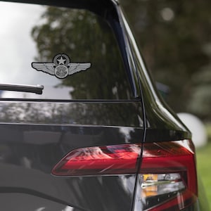 May include: A black and white sticker of a military aviation badge is affixed to the rear window of a dark gray car. The badge features a central star, surrounded by a wreath, wings, and a shield.