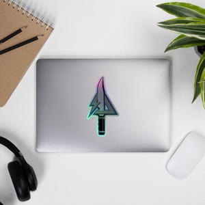May include: A sticker featuring a stylized arrow design with a gradient of pink, blue, and green. The arrow has a lightning bolt graphic and a black handle. The sticker is on a laptop, with a notebook, pencils, headphones, and a mouse nearby.