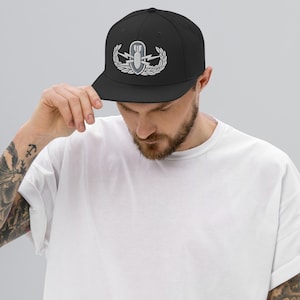 May include: A black baseball cap featuring an embroidered silver-toned military-style badge on the front. The emblem depicts a central bomb icon flanked by lightning bolts and surrounded by a wreath of laurel leaves, all rendered in shades of gray.