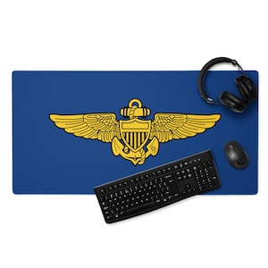 May include: A blue desk mat featuring a gold emblem with wings, an anchor, and a shield. A black keyboard, mouse, and headphones are on the mat. The mat is rectangular and designed for a computer setup.