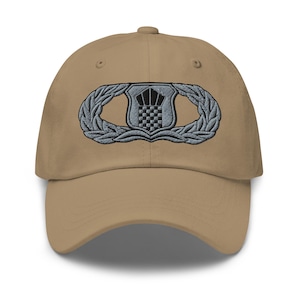 May include: A tan baseball cap with an embroidered silver and black Air Force Pararescue Jumpmaster badge.