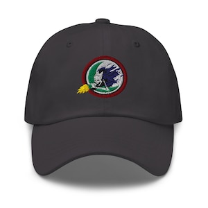 Puede incluir: Gorra de béisbol gris oscuro con un parche circular bordado. El parche presenta una calavera blanca con plumas azules y blancas, un borde verde y rojo y una llama amarilla.