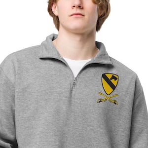 Könnte beinhalten: Ein graues Sweatshirt mit Viertelreißverschluss und einem weißen Unterhemd. Das Sweatshirt hat ein gelb-schwarzes gesticktes Emblem mit gekreuzten Schwertern. Der Kragen ist umgeklappt und der Reißverschluss ist teilweise geöffnet.