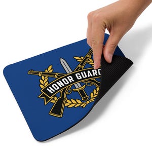 Op de afbeelding: Een rechthoekige blauwe muismat met een afbeelding van twee gekruiste geweren en een bajonet omringd door een gouden lauwerkrans. Een zwarte banner in het midden toont de witte tekst HONOR GUARD. De onderkant is van zwart rubber.