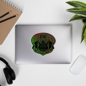 May include: A holographic sticker featuring a black graphic design with the text "JTAC" at the top and "THAT OTHERS MAY DIE" at the bottom. The design includes a globe, wings, and a hooded figure.