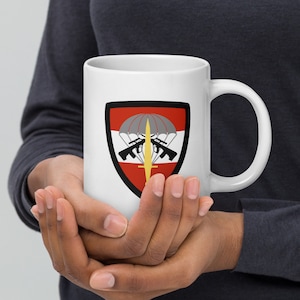 May include: White ceramic mug with a red, white, and gray military insignia featuring a parachute, crossed rifles, and a sword. The insignia is surrounded by a black border.