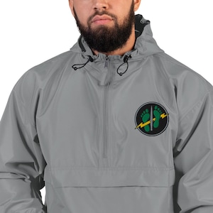 May include: A grey quarter-zip windbreaker jacket featuring a hood with black drawstrings. On the left chest, there is a circular patch displaying two green footprints, a silver dagger positioned vertically, and a yellow lightning bolt graphic.