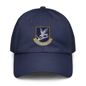 May include: A navy blue baseball cap featuring an embroidered emblem on the front. The emblem displays a white falcon perched on a branch against a blue background, enclosed in a yellow border with a banner below reading DEFENSOR FORTIS.