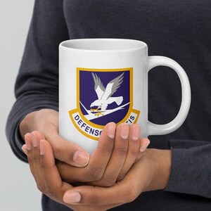 Könnte beinhalten: Eine weiße Keramiktasse mit einem blauen und goldenen Emblem, das einen weißen Vogel mit gelbem Schnabel und Krallen zeigt. Das Emblem trägt die Aufschrift "DEFENS" und "TIS".