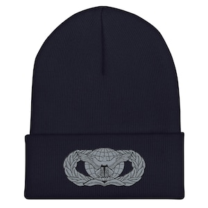 Könnte beinhalten: Marineblaue Beanie-Mütze mit silbernem Emblem. Das Emblem zeigt einen Globus mit Flügeln und einem Lorbeerkranz-Design. Die Mütze ist aus weichem Strickmaterial gefertigt.