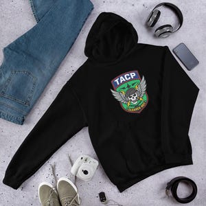 Pode incluir: Moletom preto com capuz, bolso frontal e patch gráfico. O patch apresenta uma caveira com asas, raios e o texto "TACP CLEARED HOT". O moletom é combinado com jeans azuis, tênis e acessórios.
