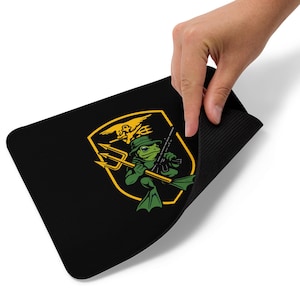 May include: Black rectangular mousepad featuring a yellow and green cartoon frog holding a trident and a rifle. The frog is wearing a hat and flippers. The design is set within a yellow shield with a gold border.