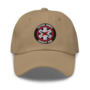 Puede incluir: Gorra de béisbol beige con visera curva. La gorra presenta un parche bordado con el texto "TACTICAL COMBAT CASUALTY CARE" que rodea un emblema blanco y azul con un rayo.