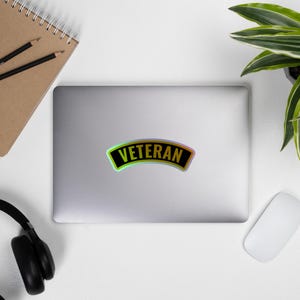 May include: A sticker with the word "VETERAN" in bold, black letters on a black background. The sticker has a curved shape with a green and yellow iridescent border. The sticker is on a silver laptop.