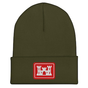 Peut inclure: Bonnet vert olive avec un écusson rectangulaire. L'écusson présente un motif de château blanc dans un cadre rouge. Le bonnet est en maille.