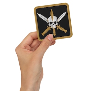 Puede incluir: Parche bordado cuadrado con fondo negro y borde dorado. El parche presenta una calavera blanca con dagas doradas cruzadas y alas. El parche se sostiene en una mano.