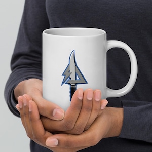 May include: White ceramic mug featuring a stylized design of a knife and arrowhead in gray, blue, and black. The mug has a large handle and is held in both hands.