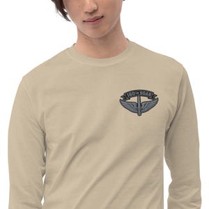 May include: A long-sleeve, tan shirt featuring a patch with the text "160th SOAR" above a winged emblem. The emblem is in shades of gray and blue. The shirt is made of a soft material.