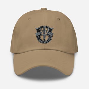 Op de afbeelding: Een beige baseballpet met een zwart en grijs geborduurd embleem. Het embleem toont een zwaard, pijlen en de tekst "DE OPPRESSO LIBER." De pet heeft een gebogen klep en een verstelbare band.