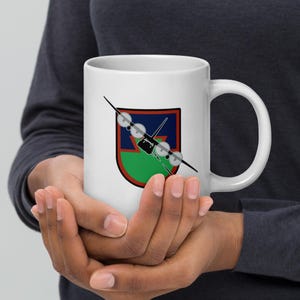 May include: White ceramic mug featuring a colorful emblem of a plane. The emblem is a shield shape with a blue and red background and a green lower section. A black plane graphic is overlaid on the shield. The mug has a large handle.
