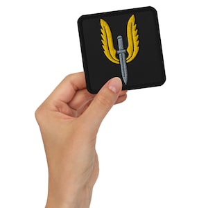 May include: Black square patch with a gold winged emblem and a silver sword. The patch is held in a hand. The design is embroidered with a black border. Ideal for jackets or bags.
