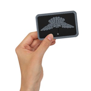 May include: A rectangular black patch with a gray border. The patch features a gray emblem of a sword with wings and a crown. The patch is held in a hand.