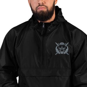 May include: A black quarter-zip windbreaker jacket featuring a hood with adjustable drawstrings. On the left chest, there is a grey graphic print depicting a scuba diving mask positioned over two crossed diving knives in a symmetrical design.