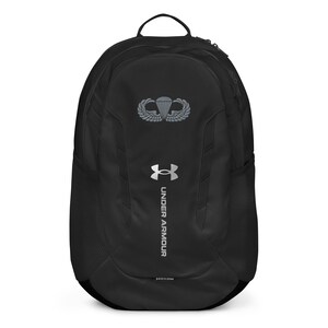 Könnte beinhalten: Schwarzer Under Armour Rucksack mit silbernem Logo und Fallschirmflügel-Emblem. Er verfügt über einen oberen Griff, seitliche Netztaschen und den Text "UNDER ARMOUR" in Silber. Der Rucksack hat ein stromlinienförmiges Design.