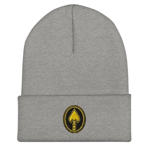 May include: A grey beanie hat with a turned-up hem. The hat has a circular emblem with a gold spade design within a black and gold rope border. The beanie is made of a soft, knitted fabric.