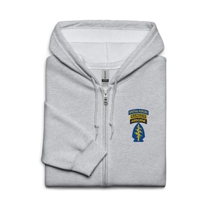 Puede incluir: Una sudadera con capucha gris claro con cremallera y capucha. La sudadera con capucha presenta un parche en el pecho con el texto "SPECIAL FORCES RANGER AIRBORNE" y un diseño de rayo. La sudadera con capucha está hecha de un material suave.