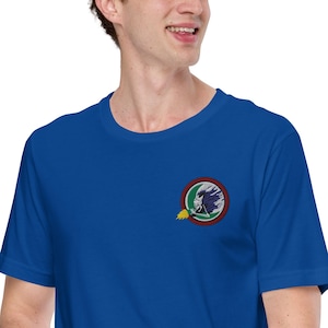 Puede incluir: Camiseta azul real con un emblema circular en el pecho izquierdo. El emblema representa una figura nativa americana con un tocado de plumas, una calavera y una llama.