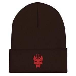 Puede incluir: Un gorro de punto marr&oacute;n con un logotipo bordado en rojo de una cabeza de animal estilizada con un tridente encima.