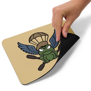 May include: A rectangular mouse pad with a tan surface featuring a cartoon green frog wearing a brown beret and parachute, with blue wings and two crossed black oars. The mouse pad has a black rubberized underside and rounded corners.
