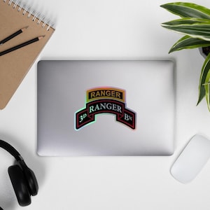 Op de afbeelding: Een sticker met de tekst "RANGER" in goud en zwart, en "3D RANGER BN" in een bannerontwerp. De sticker heeft een holografisch effect met een zwarte rand en bevindt zich op een laptop.