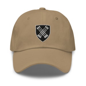 Puede incluir: Una gorra de béisbol de color beige con un escudo bordado en blanco y negro que presenta flechas cruzadas.